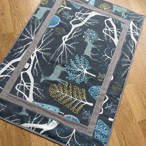 Puede incluir: Una alfombra gris y blanca con fondo azul oscuro. La alfombra presenta un patrón repetitivo de árboles estilizados, ciervos y flores. La alfombra tiene un borde gris con un borde interior blanco.