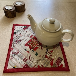 May include: A white ceramic teapot rests on a rectangular Christmas-themed placemat with a red border. The placemat features a patchwork design with festive patterns. Two small wooden candle holders with lit candles are in the background.
