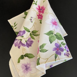 Puede incluir: Servilleta de lino crema doblada con un estampado floral. El diseño presenta flores moradas, rosas y blancas con hojas y tallos verdes. La servilleta está doblada en forma de triángulo, mostrando el estampado detallado.