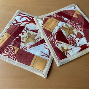 Può includere: Due presine quadrate trapuntate a tema marino. Il tessuto presenta un design patchwork con stelle marine, coralli e motivi a quadri nei toni del rosso, dell'oro e del crema. I bordi sono rifiniti con un bordo color crema.