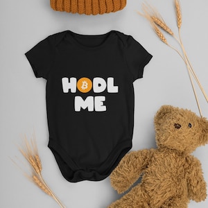 May include: Black baby onesie with white text that says "HODL ME" with a Bitcoin symbol in the middle of the word "HODL". A brown teddy bear, a brown knitted hat with a white pom-pom, and wheat stalks are also in the image.