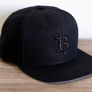Könnte beinhalten: Eine schwarze Baseballkappe mit einem schwarzen gestickten Bitcoin-Symbol auf der Vorderseite.
