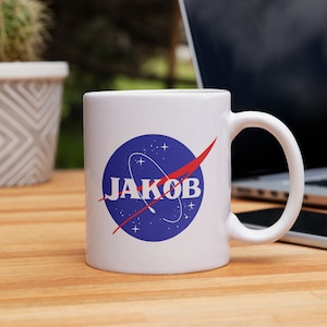 Könnte beinhalten: Weiße Keramiktasse mit einem blau-roten NASA-inspirierten Logo, auf dem "JAKOB" in weißen Buchstaben steht.