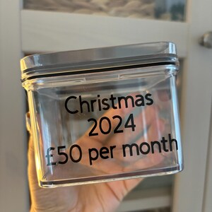Puede incluir: Un tarro de plástico transparente con tapa negra y una etiqueta que dice "Christmas 2024 £50 per month".