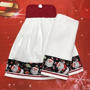 May include: A white waffle-weave kitchen towel set featuring a black border with a Santa Claus, candy cane, and snowflake print. The hanging towel has a quilted red top with a white button closure, while the second towel is folded beside it.
