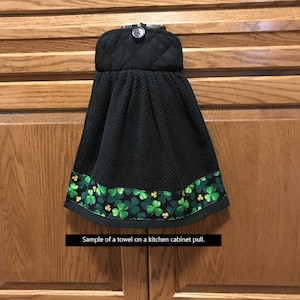 May include: Black kitchen towel with a shamrock border. The towel has a quilted top with a button closure. The bottom edge features a green and gold shamrock pattern on a black background. The text on the image says "Sample of a towel on a kitchen cabinet pull."