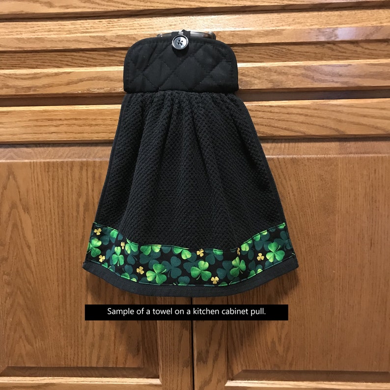 May include: Black kitchen towel with a shamrock border. The towel has a quilted top with a button closure. The bottom edge features a green and gold shamrock pattern on a black background. The text on the image says "Sample of a towel on a kitchen cabinet pull."
