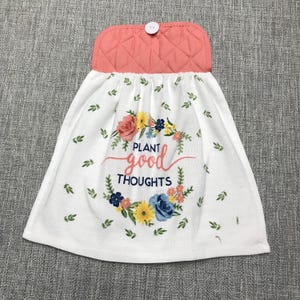 May include: White kitchen towel with a floral wreath design and the words "Plant Good Thoughts." The towel has a coral-colored quilted top with a button closure. Green leaves and colorful flowers decorate the towel.