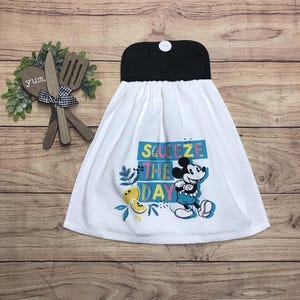 May include: White kitchen towel with a black top and a button closure. The towel features a colorful graphic of Mickey Mouse and the phrase "Squeeze the Day". Decorative wooden utensils and greenery are in the background.