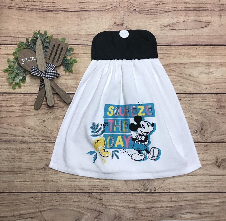 May include: White kitchen towel with a black top and a button closure. The towel features a colorful graphic of Mickey Mouse and the phrase "Squeeze the Day". Decorative wooden utensils and greenery are in the background.