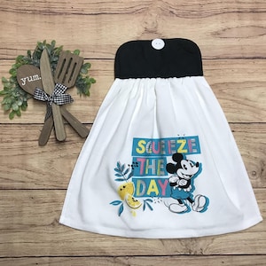 May include: White kitchen towel with a black top and a button closure. The towel features a colorful graphic of Mickey Mouse with the words "Squeeze the Day". Decorative lemons and leaves are also included in the design.