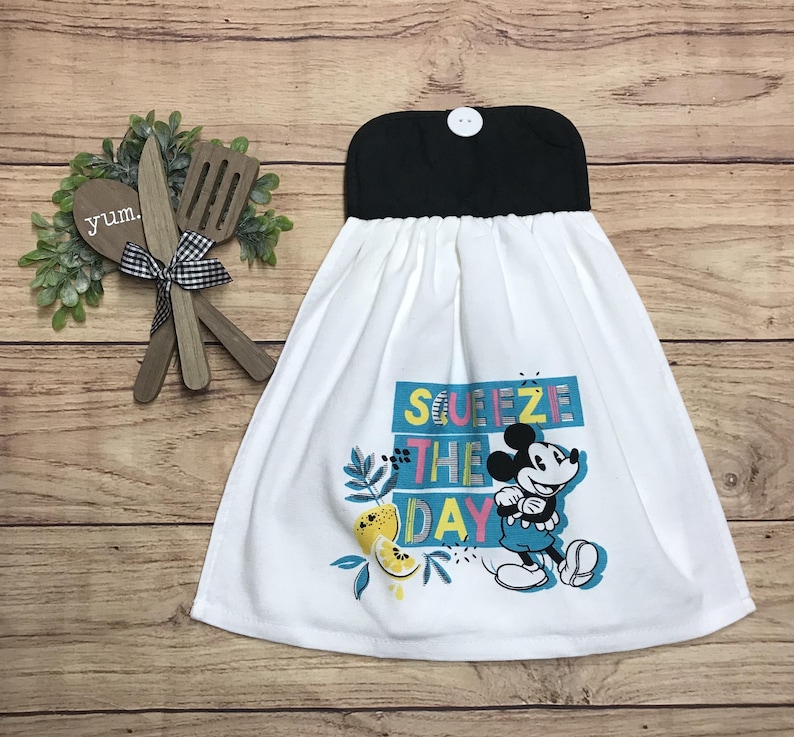 May include: White kitchen towel with a black top and a button closure. The towel features a colorful graphic of Mickey Mouse with the words "Squeeze the Day". Decorative lemons and leaves are also included in the design.