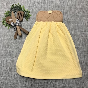 May include: Yellow kitchen towel with a quilted tan top section and a button closure. Wooden utensils and a decorative sprig of greenery are to the left of the towel. The word "yum" is on a wooden spoon.
