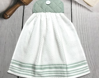 May include: A white kitchen towel with a textured weave and a light green striped border. The top of the towel has a quilted light green section with a button. A wooden rolling pin is in the background.
