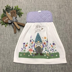 May include: White kitchen towel with a gnome design and the words "let love grow." The gnome wears a blue hat and is surrounded by colorful flowers and mushrooms. The top of the towel is lavender and quilted.