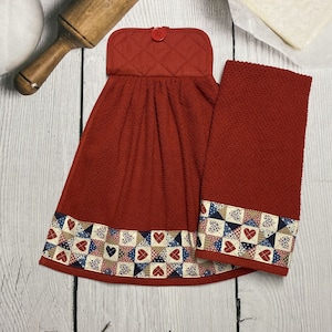 May include: A set of two red kitchen towels. One is a hanging towel with a quilted top and a button closure. Both towels have a decorative border with heart and star patterns in red, white, and blue.
