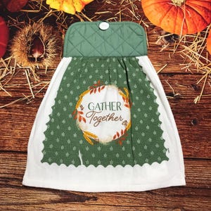 May include: A hanging kitchen towel featuring a quilted green top with a white button closure. The main fabric is green with a small white leaf pattern and a central white circular graphic containing the text GATHER Together.