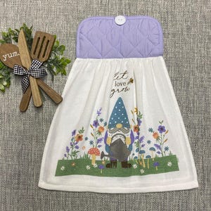 May include: A white kitchen towel with a lavender quilted top and a button closure. The towel features a gnome design with the words "let love grow" and colorful flowers. Wooden utensils and greenery are to the left.