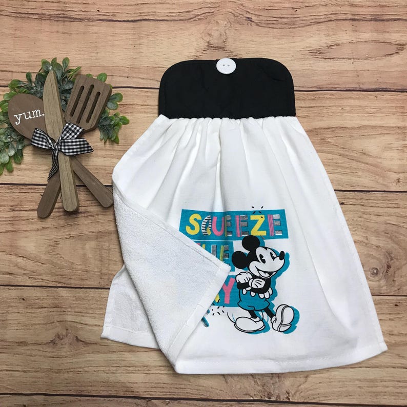 May include: White kitchen towel with a black top and a button closure. The towel features a colorful graphic of Mickey Mouse with the text "Squeeze the Day". The towel is hanging on a wooden surface.