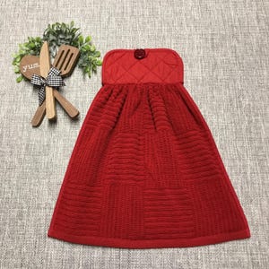 May include: A red kitchen towel with a quilted top and a button closure. The towel has a textured pattern and is hanging. A wooden decorative piece with the word "yum" and kitchen utensils is to the left.