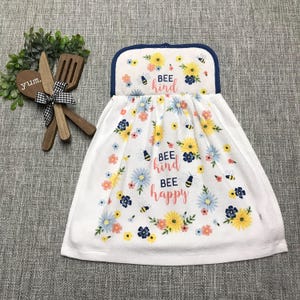 May include: A white hanging kitchen towel featuring a floral pattern with yellow, blue, and pink flowers, small bees, and the text "BEE kind BEE happy" in blue and pink script. Beside it are two wooden kitchen utensils tied with a black and white gingham bow.