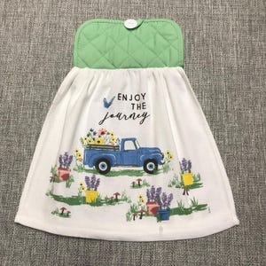 May include: White kitchen towel with a green quilted top and a button closure. The towel features a blue truck carrying flowers, with the text "Enjoy the Journey". Colorful potted plants and mushrooms are also depicted.