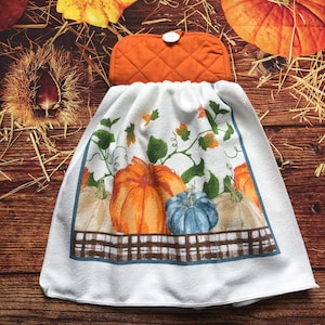 May include: A white hanging kitchen towel featuring a printed design of orange, blue, and cream pumpkins with green vines and leaves, finished with a brown and white plaid border and a quilted orange fabric top with a white button closure.
