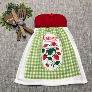 May include: A hanging kitchen towel featuring a green and white gingham pattern with a central graphic of strawberries and the text Welcome TO OUR PATCH. The towel has a red quilted fabric top with a red button and is paired with wooden utensils.