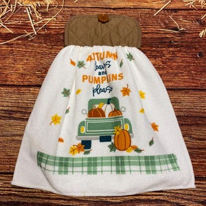 May include: A white hanging kitchen towel with a quilted brown top and a single button. The towel features a green truck graphic carrying pumpkins, scattered autumn leaves, and the text "AUTUMN leaves and PUMPKINS please" in orange and blue.