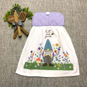 May include: A white hanging kitchen towel with a quilted lavender top and a button closure. The towel features a graphic of a gnome in a blue hat holding a watering can, surrounded by colorful flowers and mushrooms, with the text let love grow.