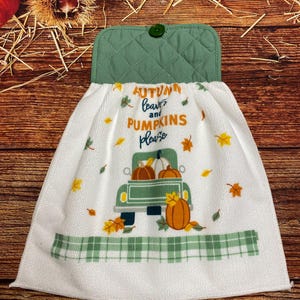 May include: A white hanging kitchen towel with a green quilted top and a single green button. The towel features a graphic of a green truck carrying orange pumpkins, scattered autumn leaves, and the text "AUTUMN leaves and PUMPKINS please."
