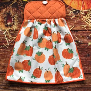 May include: A hanging kitchen towel featuring a white background with an all-over print of orange pumpkins and green leaves. The towel has a quilted orange fabric top section with a white button closure, and an orange fabric border trim.