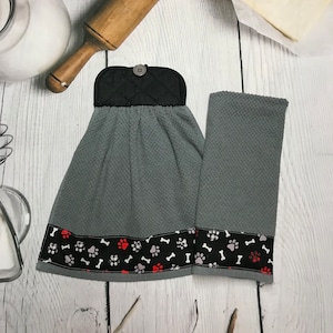 May include: Gray kitchen towel set with a black and white paw print and bone pattern border. The hanging towel has a black quilted top with a button. The set includes a matching hand towel.