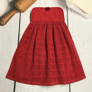 May include: A red kitchen hand towel with a quilted top and a button closure. The towel has a textured, checkered pattern and is hanging on a white wooden surface. A rolling pin and other kitchen items are in the background.