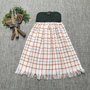 May include: A kitchen towel with a green quilted top and a button closure. The towel features a white background with an orange and gray plaid pattern and a fringed bottom edge. Decorative wooden utensils and the word "yum" are also visible.