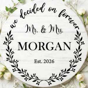 May include: A white wooden round sign with the text "we decided on forever Mr. & Mrs. MORGAN Est. 2026" in black. The sign is surrounded by a black floral wreath and white flowers.