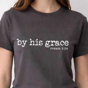 May include: Dark gray t-shirt with the text "by his grace" in white, along with the reference "romans 3:24". The shirt has short sleeves and a crew neck.