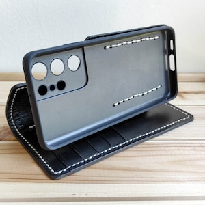 Puede incluir: Funda de teléfono de cuero negro con función de soporte. La funda tiene costuras blancas y un recorte para el objetivo de la cámara.