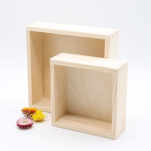 Peut inclure: Deux boîtes en bois non finies de tailles différentes, disposées côte à côte. Les boîtes sont rectangulaires avec des faces avant et arrière ouvertes. Quelques fleurs séchées colorées sont à la base des boîtes. Les boîtes sont en bois clair.