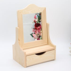 Può includere: Uno specchio da toeletta in legno chiaro con un cassetto. Lo specchio ha una parte superiore ad arco ed è posto su una base rettangolare. Lo specchio riflette una colorata composizione floreale. Il cassetto ha una maniglia curva.