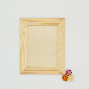 Puede incluir: Un marco de fotos de madera natural con un acabado marrón claro. El marco está vacío y listo para una foto o una obra de arte. Tres flores secas se encuentran en la esquina inferior derecha de la imagen.