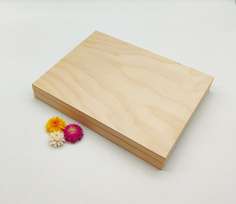 Pode incluir: Uma caixa de madeira de cor clara com tampa, sobre um fundo branco. A caixa &eacute; retangular e parece ser feita de madeira natural. Um pequeno grupo de flores secas coloridas em amarelo, laranja, branco e rosa &eacute; colocado perto da caixa.
