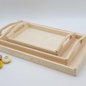 Può includere: Tre vassoi in legno con manici, uno dentro l'altro. I vassoi sono in legno naturale e hanno un colore marrone chiaro.