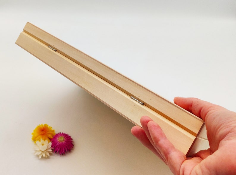 Pode incluir: Uma moldura de madeira clara, segurada em &acirc;ngulo, com uma dobradi&ccedil;a vis&iacute;vel. A moldura &eacute; feita de duas pe&ccedil;as de madeira conectadas. Tr&ecirc;s flores secas em amarelo, branco e rosa est&atilde;o no canto inferior esquerdo.