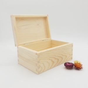 May include: An open, rectangular wooden box with a hinged lid. The box is made of light-colored wood and features dovetail joints at the corners. Two dried flowers in shades of purple and yellow are next to the box. The box is empty.