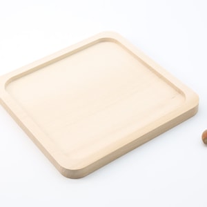 Könnte beinhalten: Ein quadratisches, helles Holztablett mit abgerundeten Ecken. Das Tablett hat eine vertiefte Mitte und ist von einem erhöhten Rand umgeben. Eine Walnuss und zwei Haselnüsse liegen neben dem Tablett.