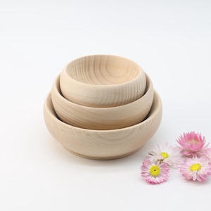 May include: Three wooden bowls of varying sizes are stacked on top of each other. The bowls are made of light-colored wood and have a smooth, rounded shape. Pink flowers are scattered on the white surface below the bowls.