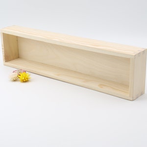 Może przedstawiać: Prostokątna, niewykończona drewniana półka z pustym wnętrzem. Półka wykonana jest z jasnego drewna i ma prosty, minimalistyczny design. Mała wiązka żółtych, różowych i białych suszonych kwiatów leży w pobliżu półki.