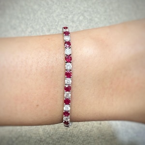 Peut inclure: Un bracelet de tennis avec des pierres précieuses rouges et claires alternées, serties dans un bracelet argenté. Le bracelet est porté au poignet, mettant en valeur le design symétrique et les couleurs vives des pierres.
