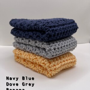 May include: A stack of three crocheted dishcloths in navy blue, dove grey, and banana yellow.